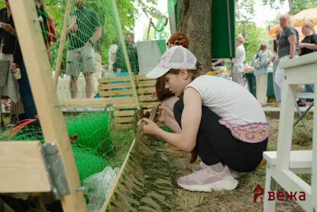 Girl kneeling by a green chicken-wire fence, tying brown strips of material during an outdoor event; spectators in background.