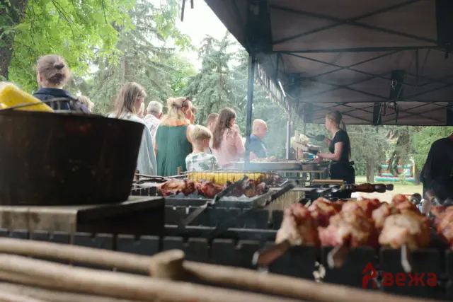 Outdoor barbecue scene under a canopy with people lined up and a cook grilling meat on a smoky grill.