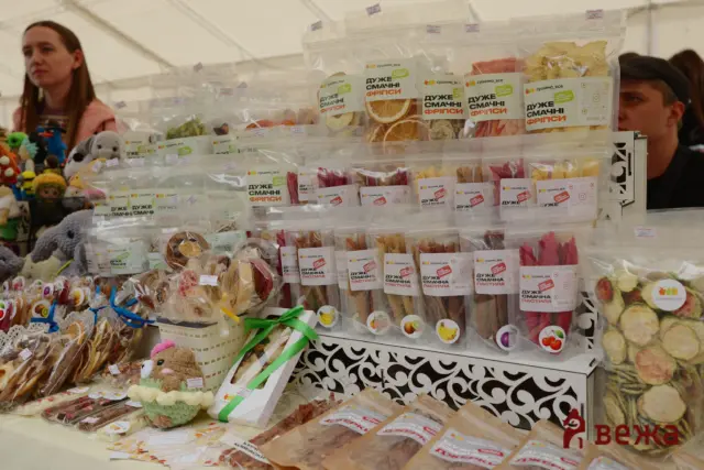 Display of packaged dried snacks in clear bags with Cyrillic labels at a market stall; a woman is in the background.