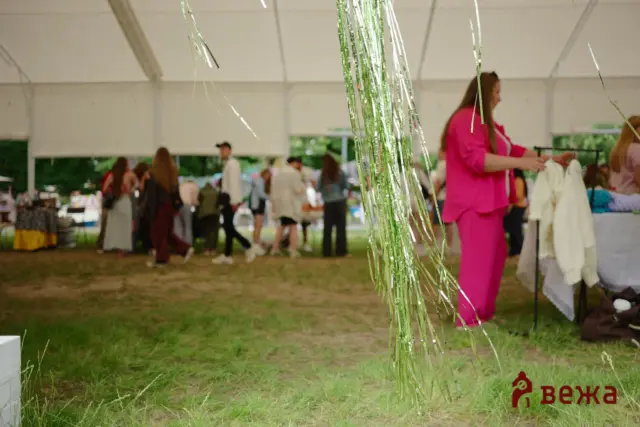 Outdoor event under a white tent with metallic streamers in the foreground and people browsing stalls; a person in pink dress on the right side.