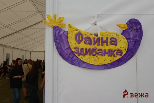 Colorful bird-shaped sign in purple and yellow with Cyrillic text, mounted on a white wall at an indoor event venue.
