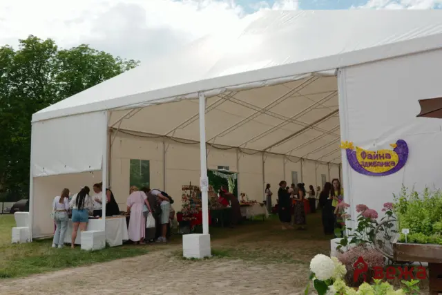Outdoor festival booth under a white canopy with people browsing tables of crafts; trees in the background.