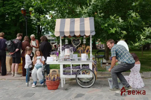 Outdoor market stall selling lavender products under a striped beige-gray canopy, with people browsing nearby.