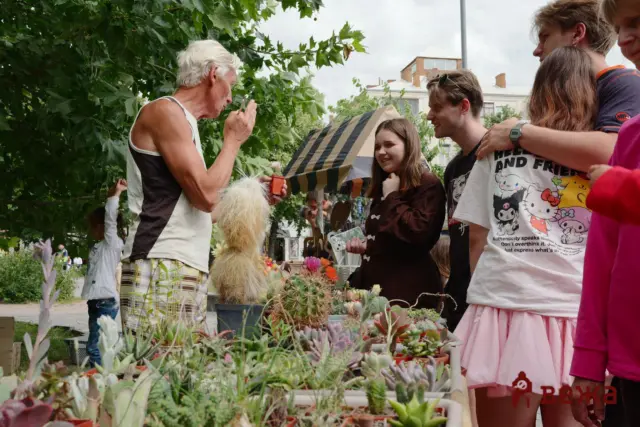 Older man with white hair showing a small potted plant to smiling young people at an outdoor market with succulent plants in the foreground.