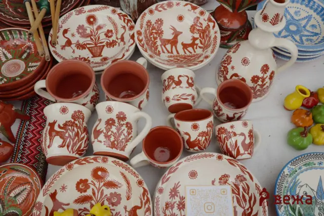 Display of white pottery with red folkloric floral and animal designs, including plates, cups, and a pitcher, all arranged together.