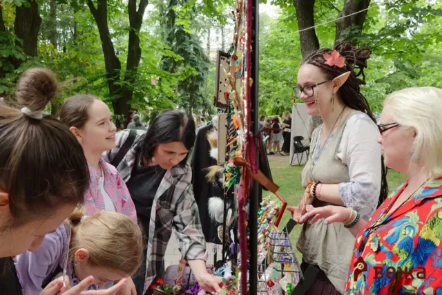 People gathered at an outdoor market browsing a jewelry display with colorful beaded accessories; a vendor wearing elf ears and a maple leaf in her hair smiles nearby.