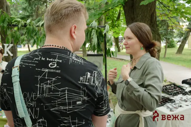 Two adults chat in a park: a man with a graphic black shirt and teal bag facing a woman in a green jacket near a booth.