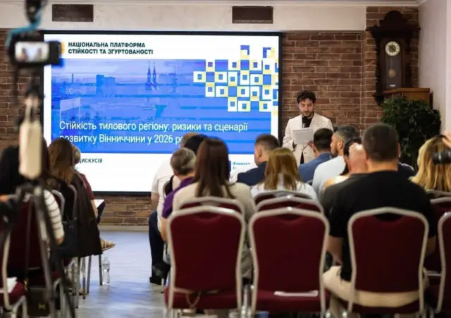 Presenter standing at a lectern presenting to a seated audience in a brick-walled conference room; a large blue slide with Ukrainian text is displayed behind him.