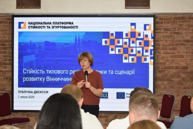 Woman in a maroon top speaks into a microphone in front of a large blue presentation slide with Ukrainian text and orange–blue squares edge design.
