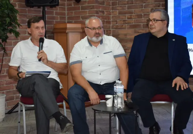 Three men sit on stage for a panel discussion in front of a brick wall; the left speaker holds a microphone and papers.