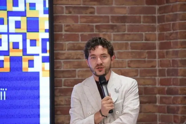 Man in a light blazer speaking into a handheld microphone in front of a brick wall backdrop.