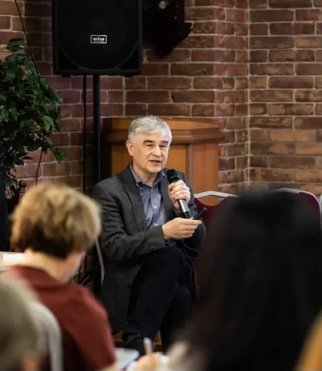 Man in a gray blazer speaks into a handheld microphone during a talk, seated in front of a brick wall with an audience nearby.