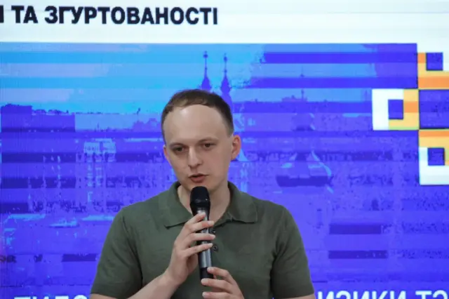 Young man in a green polo shirt speaks into a handheld microphone against a blue, pixelated backdrop.