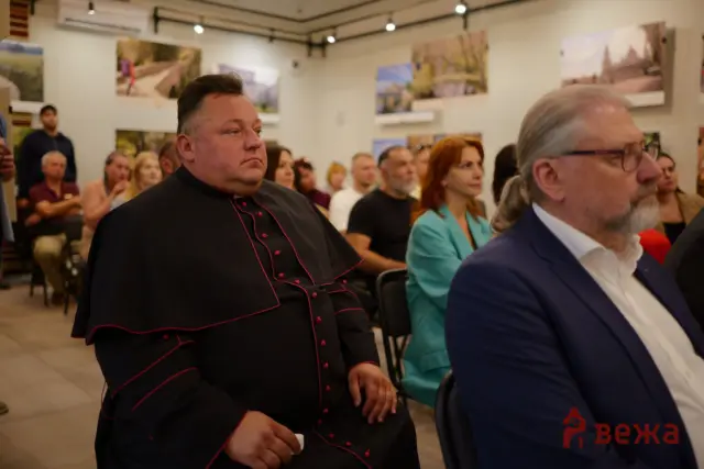 Man in black clerical-style outfit with red piping seated in foreground at an indoor event, with a crowd listening in a gallery-like room.
