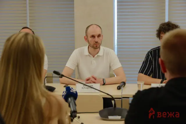 Man in white polo speaking at a panel table with microphones, audience members in foreground in a conference room.