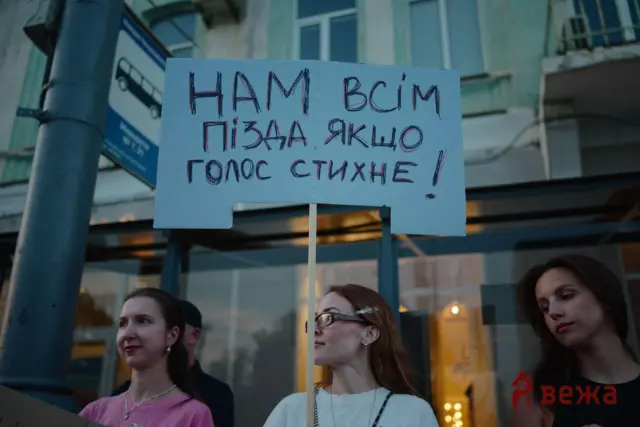 Group of people outdoors holding a light-blue cardboard sign with large handwritten Cyrillic text.