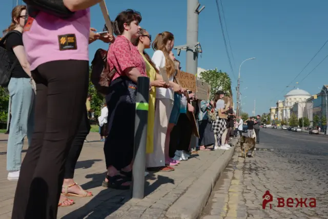 Group of people standing along a sidewalk at a protest, holding cardboard signs on a sunny day, with a dog nearby on the street.