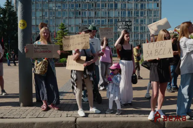 Group of protesters holding handwritten cardboard signs in Ukrainian, standing on a paved square with a glass-front building in the background; a child drinks from a pink bottle in the foreground.