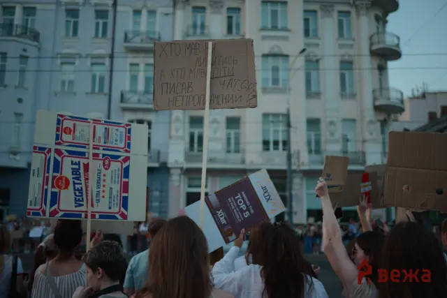 Crowd of protesters holding cardboard signs with Cyrillic text in a city street, with apartment buildings in the background.