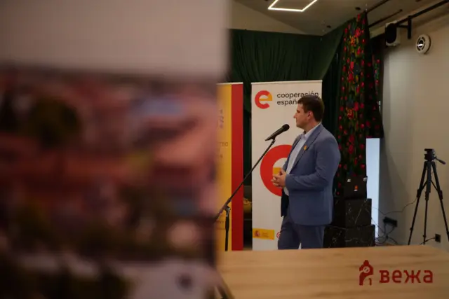 Speaker in a blue suit addresses the audience from a podium on stage, with a 'cooperación española' banner behind him and a floral curtain nearby.