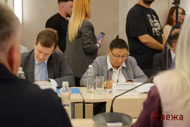 Conference table with attendees signing documents; name placards, water bottles, and a camera stand visible in the background.