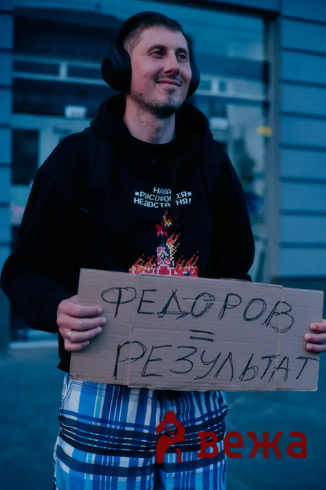 Man wearing a black hoodie and large headphones holds a cardboard protest sign with handwritten Cyrillic text, outdoors against a building.