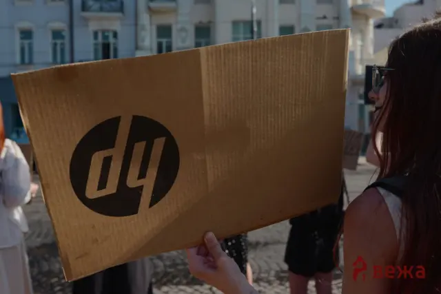 Person holds up a large cardboard sign with a bold black circular logo, partially covering their torso on a sunny city street, with buildings in the background.