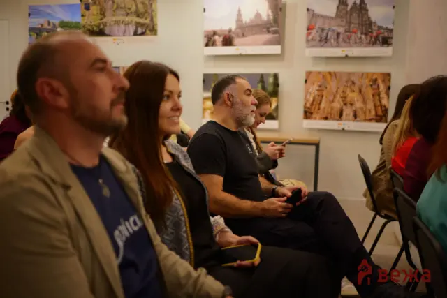 Audience of adults seated in a room, facing forward as a presentation presumably takes place; travel photos hang on the wall behind them.