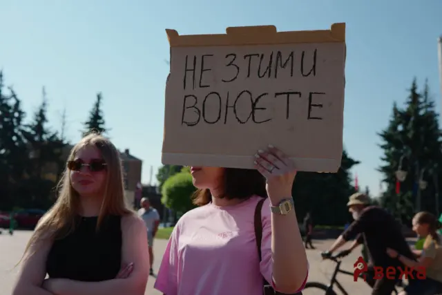 Person holding a cardboard protest sign with large handwritten letters, outdoors with trees and people in the background (sign is partially legible).