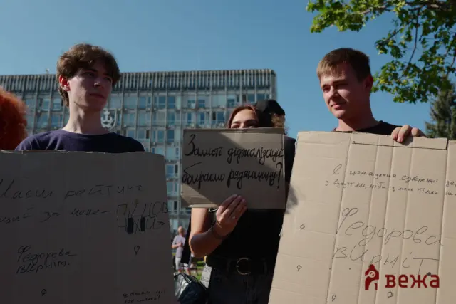 Group of people holding large cardboard signs in a sunny outdoor protest scene, with a building and trees in the background.