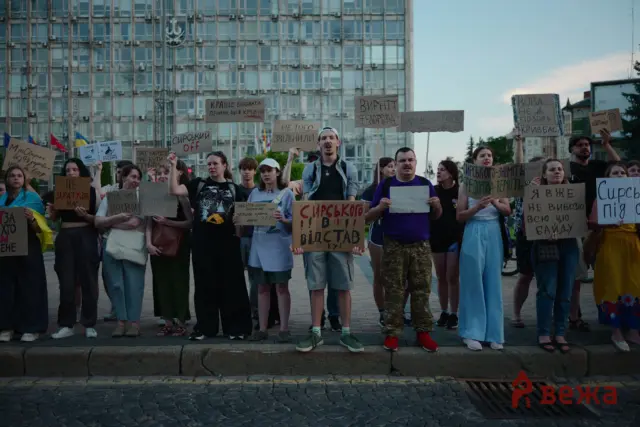 Group of protesters in a city square holding handwritten signs in Cyrillic, with a glass-front building in the background and a few flags visible on the left.