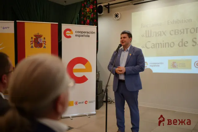 Man in a blue suit giving a talk on stage with banners showing the Spanish flag and 'cooperación española' logos.