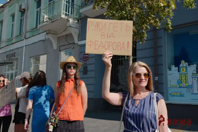 Protesters on a city street, a cardboard sign reading 'РУКИ ГЕТЬ ВІД ФЕОДОРОВА!' held up in the foreground by a smiling woman in sunglasses.