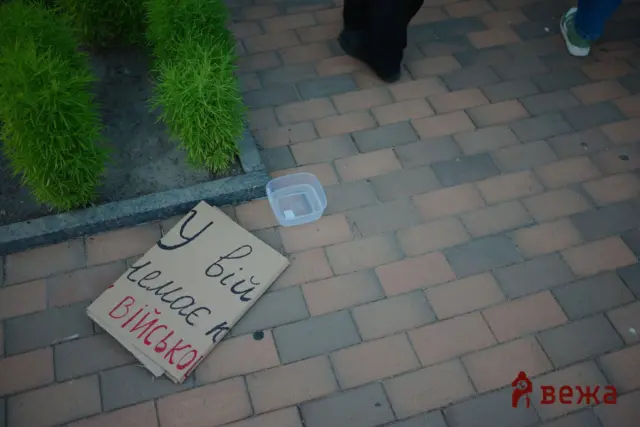 Cardboard sign with handwritten Cyrillic text on a brick sidewalk, next to a small clear plastic container, with greenery on the left and pedestrians walking by.