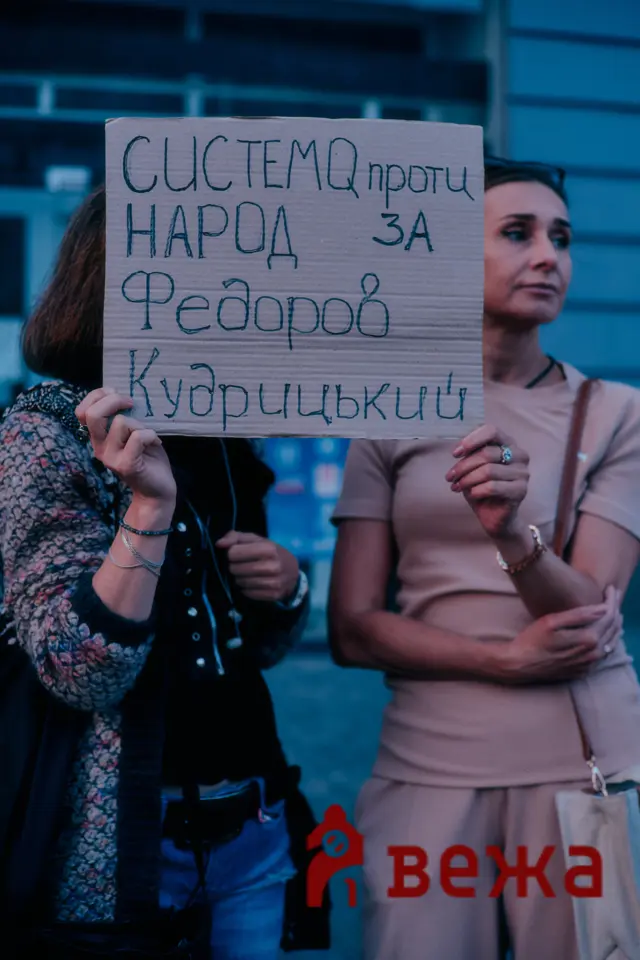 Two women stand at a protest, one holding a cardboard sign with handwritten Cyrillic text.