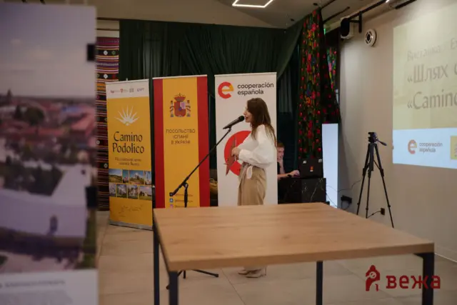 Woman in a white blouse speaks at a microphone in a conference room with banners for Spain and cooperation española behind her.