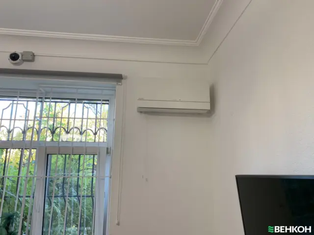 White wall-mounted air conditioner above a window with metal bars; a security camera sits near the ceiling on the left.