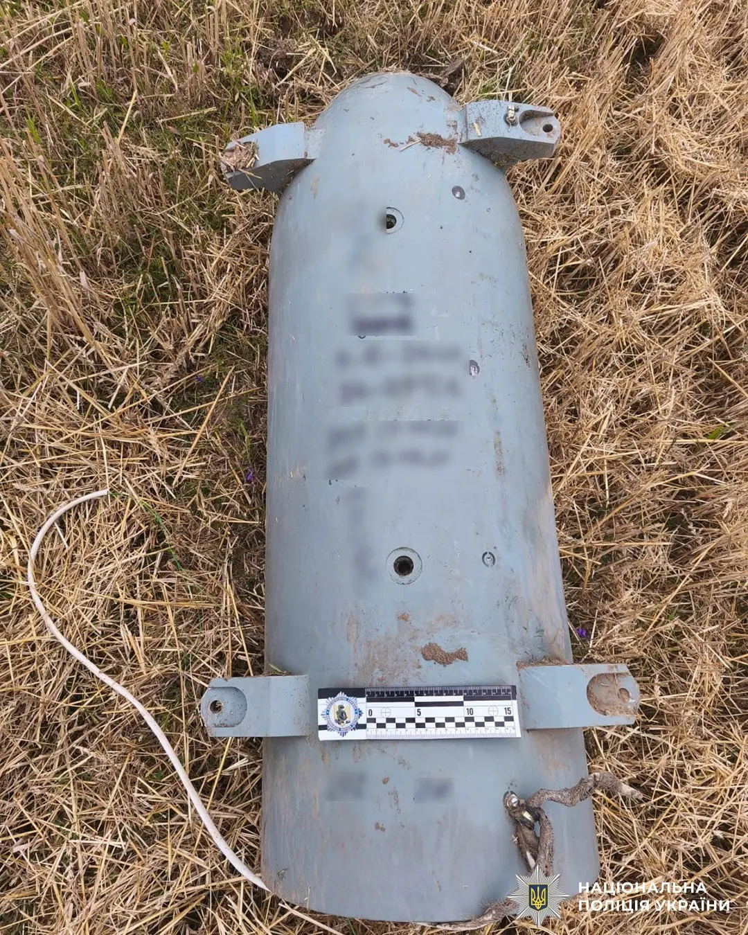 Large blue-gray cylindrical munition casing with mounting brackets lying in dry grass, next to a measurement scale and police emblem nearby.