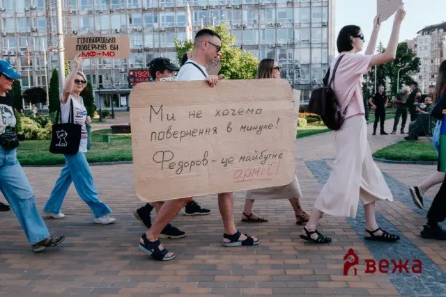 Group of protesters walking in a plaza, carrying large cardboard signs with handwritten Cyrillic text; urban background with a glass-fronted building.