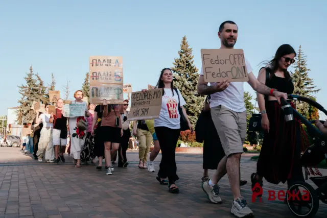 Group of protesters walking on a sunny street, holding large cardboard signs with handwritten messages.