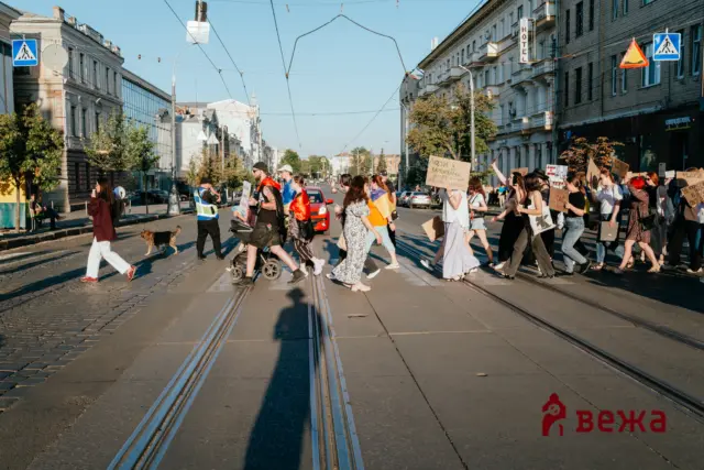 A group of protesters crosses a city street on tram tracks, carrying cardboard signs; some wear orange and blue/yellow flags, with tall buildings along the avenue and a clear sky.