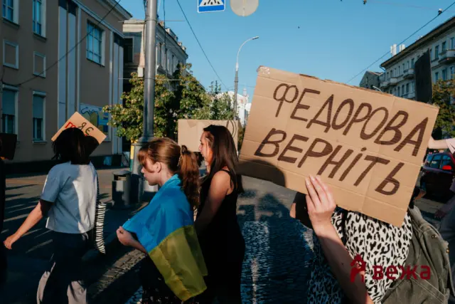 Protesters walk down a sunny city street, holding large cardboard signs with Cyrillic text; a blue-yellow flag draped over one protester’s shoulders.