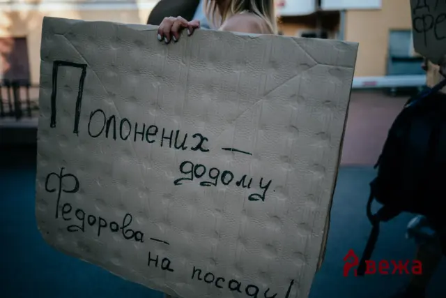 Person holding a large, hand-painted cardboard sign with Cyrillic writing outdoors, partially obscuring their face.