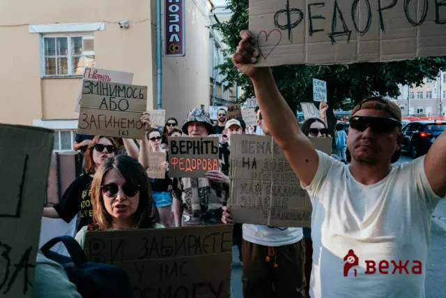 Crowd of protesters holding handwritten cardboard signs in Cyrillic, marching on a city street; a man in a white shirt holds an overhead sign, storefronts and trees in the background.