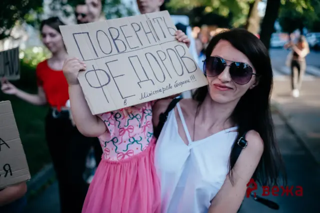 Woman with dark hair and sunglasses holds a cardboard protest sign with large handwritten Cyrillic text at a street demonstration, crowd blurred in the background.