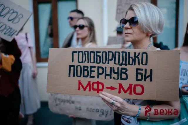 Protester holding a cardboard sign with bold Cyrillic text at a street demonstration.