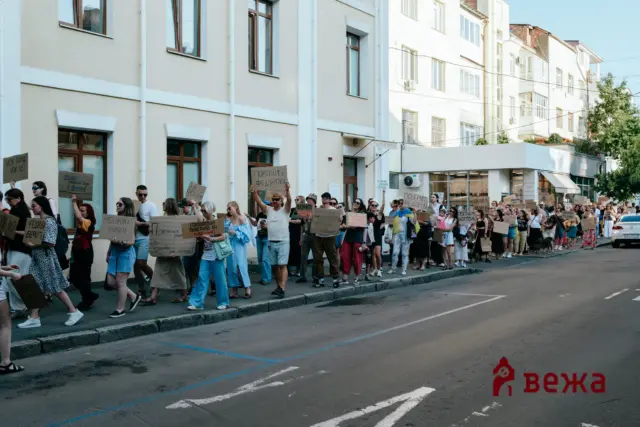 Crowd of protesters walking down a city street, holding handmade cardboard signs, with light-colored apartment buildings in the background.