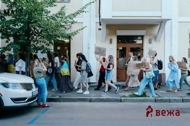 Group of protesters walking along a city street, holding cardboard signs as they pass a beige building with wooden doors.