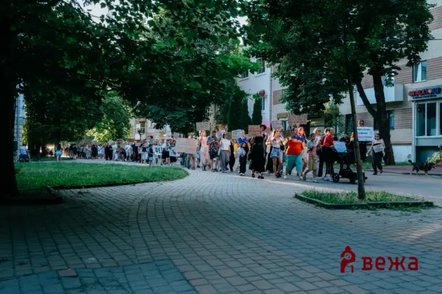 Crowd of protesters marching along a tree-lined urban street, carrying handmade signs.