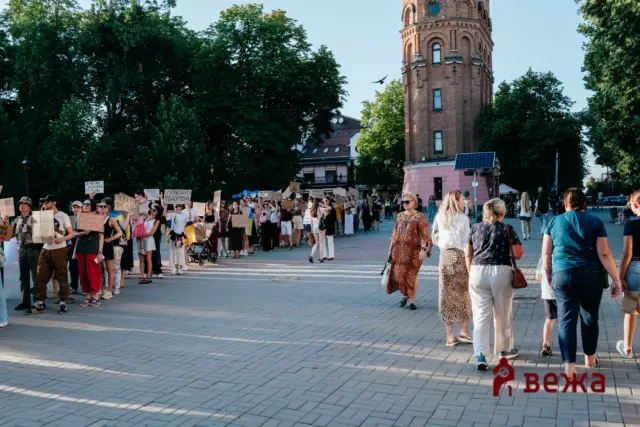 Crowd of protesters holding handwritten signs in a city square, with a tall brick tower and trees behind.